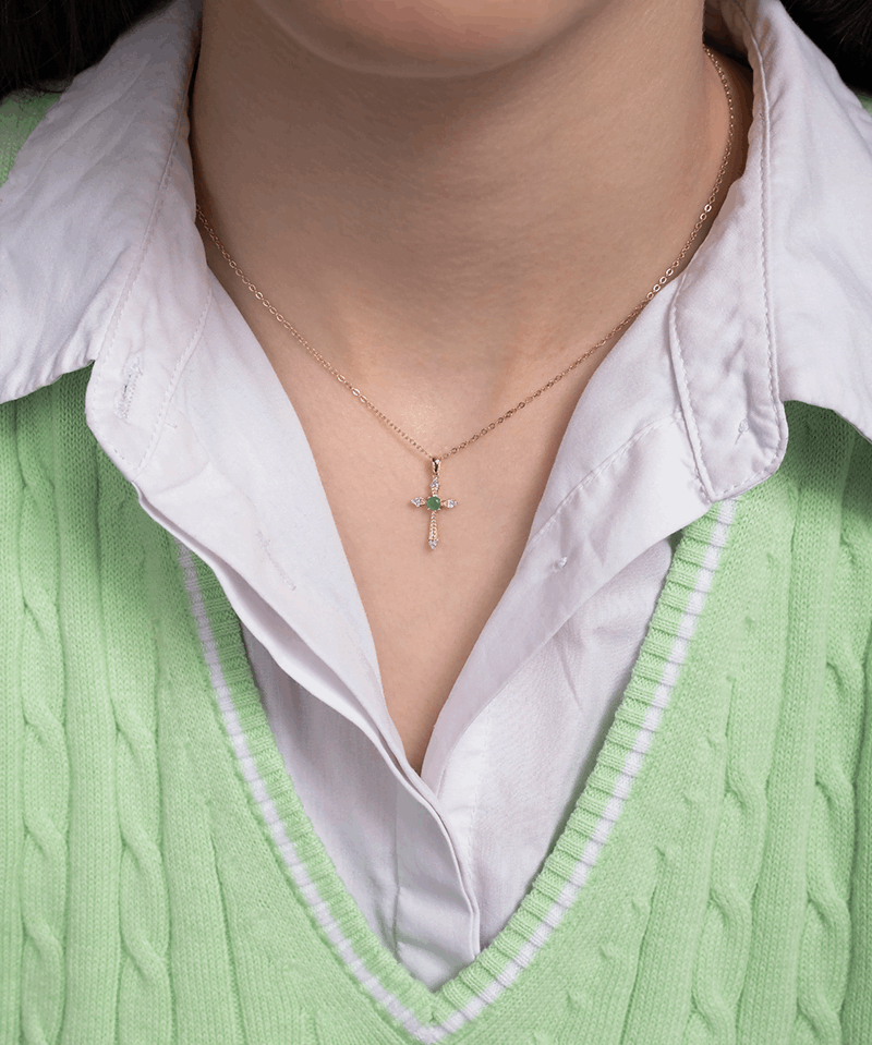 Wisiorek krzyż ze szmaragdem na delikatnym łańcuszku, noszony na szyi przez osobę w zielonym swetrze