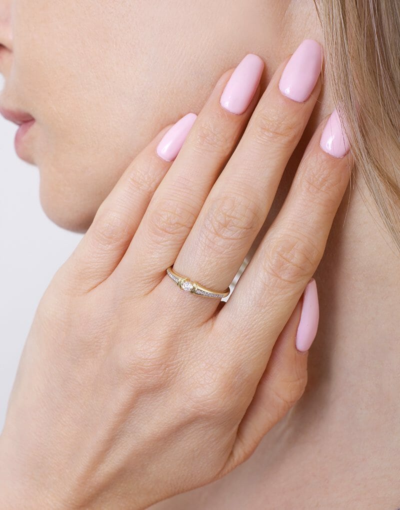 Kobieta nosi złoty pierścionek z pół-paskami brylantów na palcu, z różowym manicure
