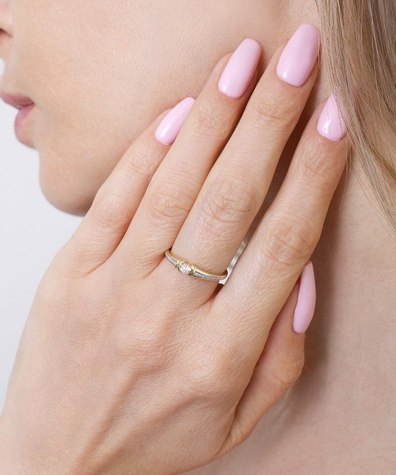 Kobieta nosi złoty pierścionek z pół-paskami brylantów na palcu, z różowym manicure